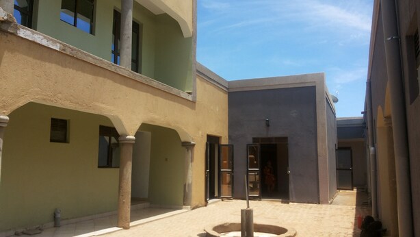 A courtyard with a fountain, a building with a grey facade, and a green building with a balcony. - Skypa Hotel (Yumbe)