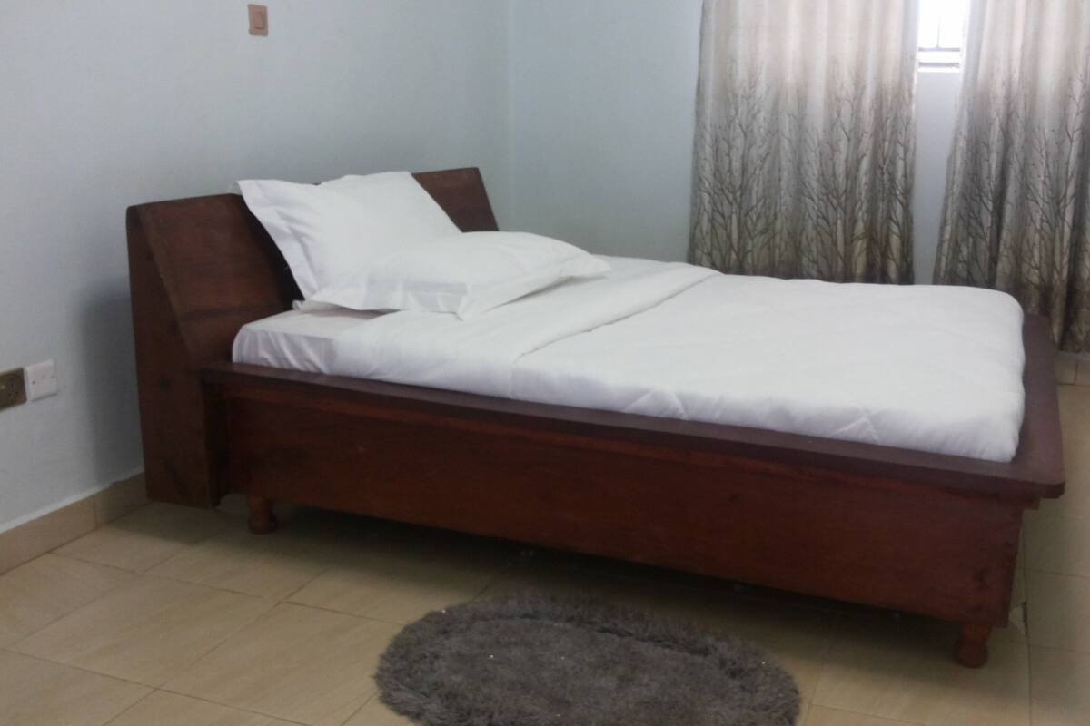 A single bed with a wooden frame and white bedding in a room with light-colored walls and a tiled floor.