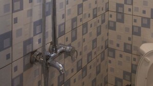 A shower with a handheld showerhead and a wall-mounted rain showerhead. The shower is installed in a corner with tiled walls featuring a geometric pattern. - Skypa Hotel (Yumbe)