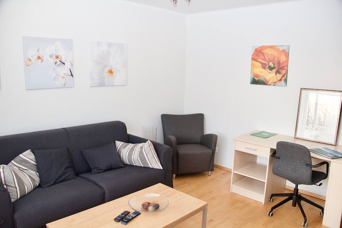 A modern living room with a dark grey sofa, a grey armchair, a wooden coffee table with a bowl and remote controls, a desk with a chair, and three framed artworks on the walls.