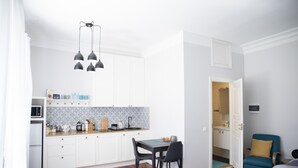 Cocina compacta con muebles blancos, un microondas y un salpicadero con estampado. Hay una mesa de comedor con sillas, un tapete azul y una silla azul en la sala.