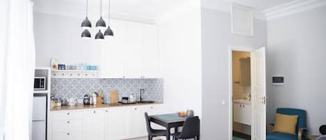 A compact kitchen with white cabinets, a microwave, and a patterned backsplash. There is a dining table with chairs, a blue rug, and a blue chair in the living area.