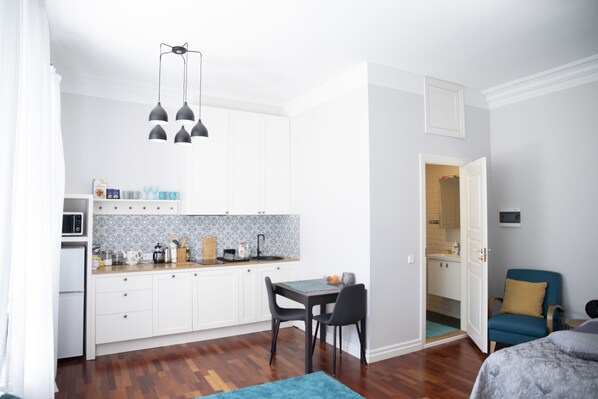 A compact kitchen with white cabinets, a microwave, and a patterned backsplash. There is a dining table with chairs, a blue rug, and a blue chair in the living area. - Old town Library Tartu Home Apartments (Tartu)