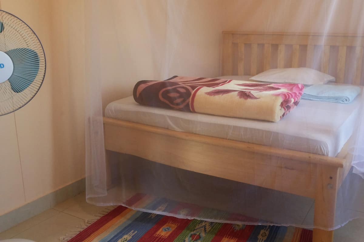 Una cama individual con cabecera de madera, un colchón blanco y una manta con estampado floral. Un mosquitero cubre la cama, y un ventilador está montado en la pared a la izquierda.
