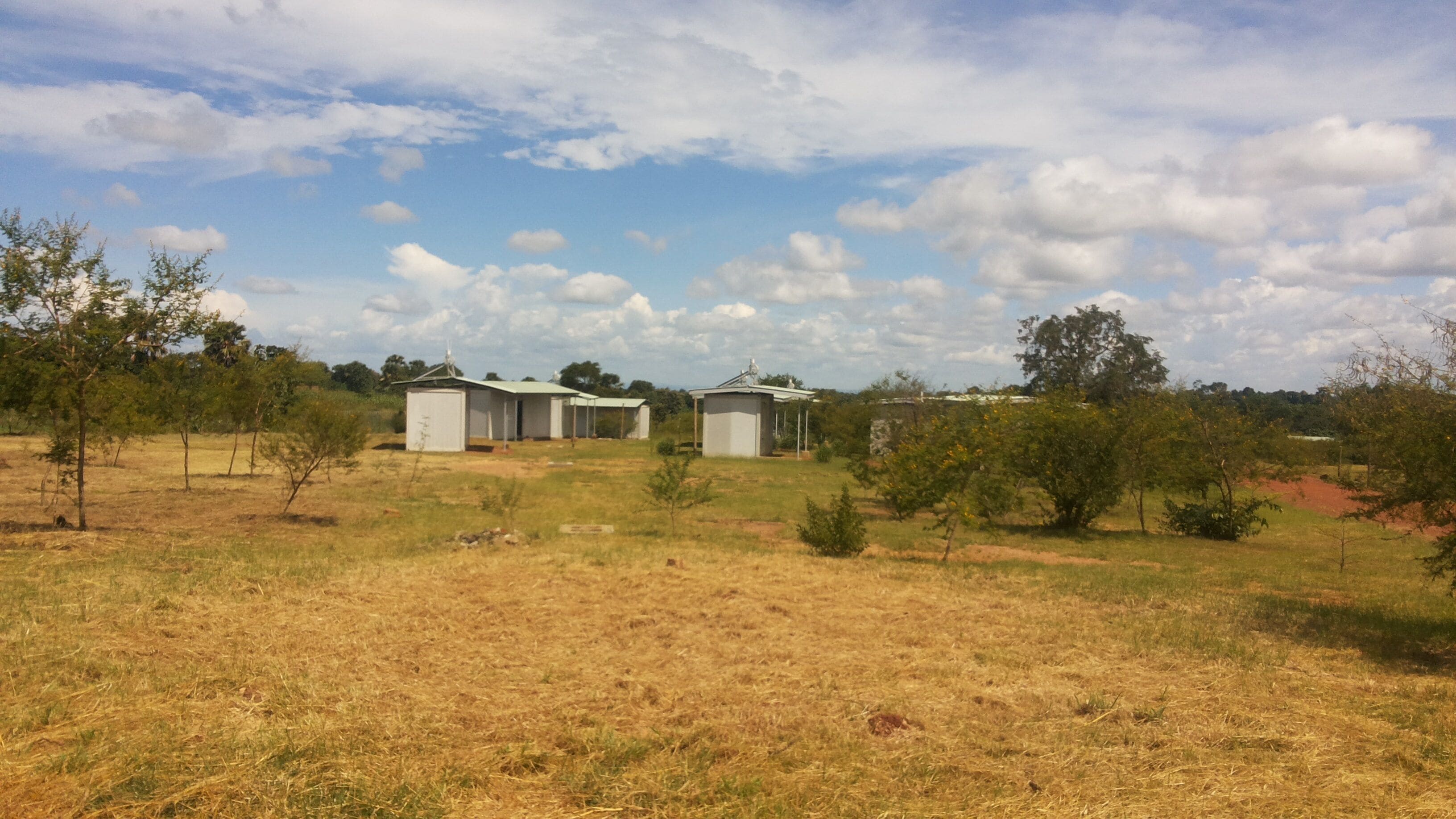 Entorno rural con tres pequeñas construcciones blancas de un piso y techos planos, rodeado de hierba seca y escasos árboles.