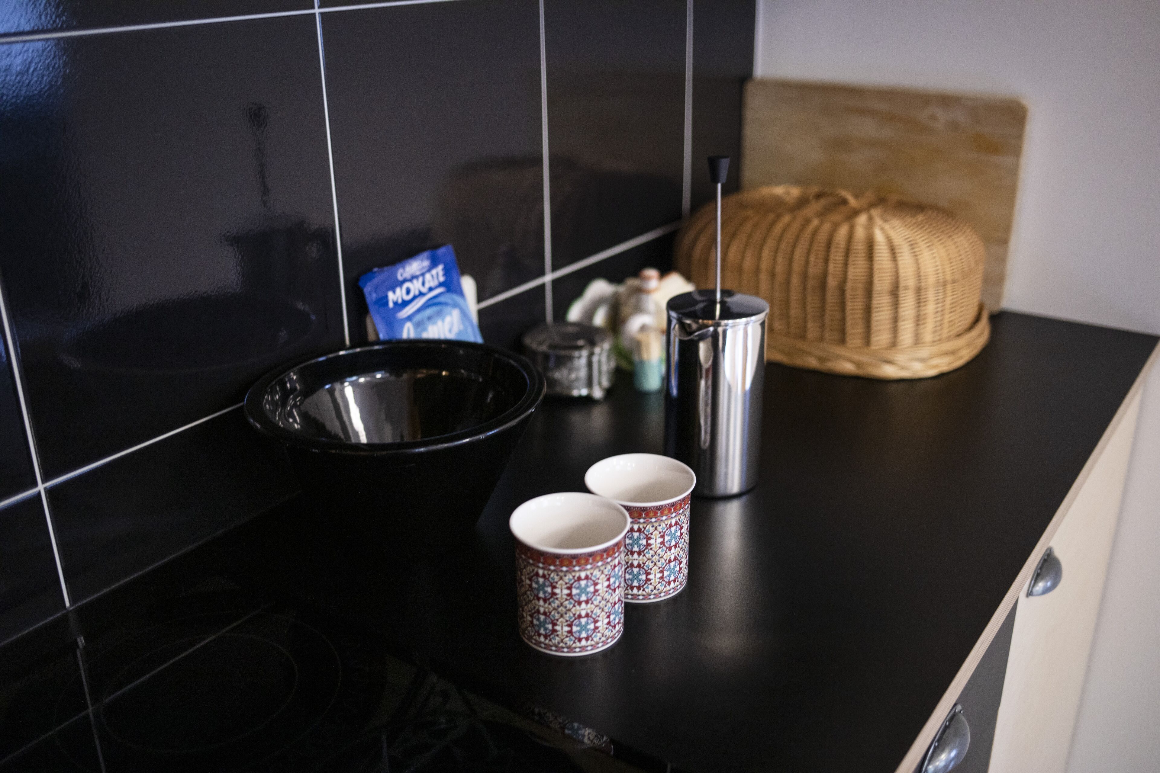 A kitchen counter with a black bowl, two patterned cups, a metal container, and a woven basket.