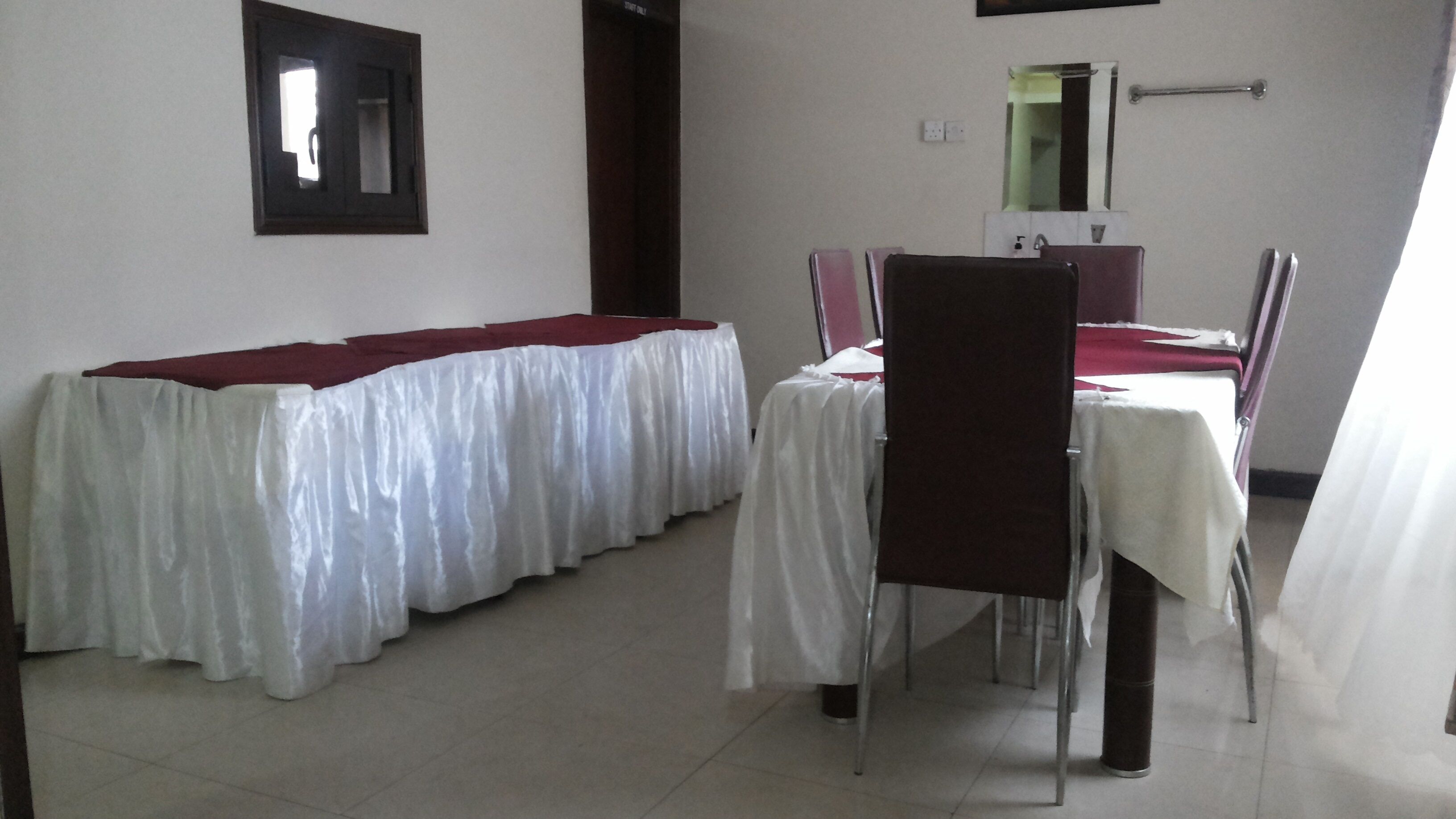 Habitación de hotel con comedor que cuenta con dos mesas, cada una con mantel blanco y camino de mesa rojo. Hay un espejo en la pared sobre el comedor y una ventana con vista al exterior.