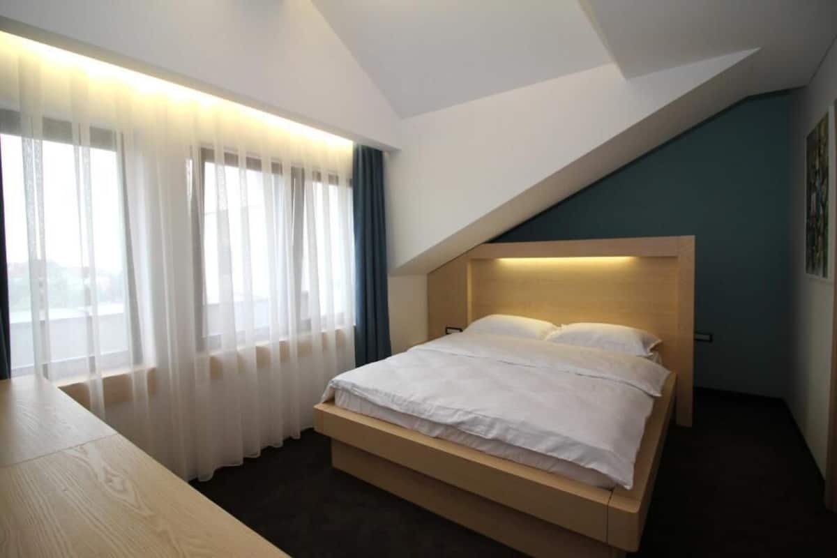 A modern hotel room with a wooden bed, a desk, and a large window with sheer curtains.