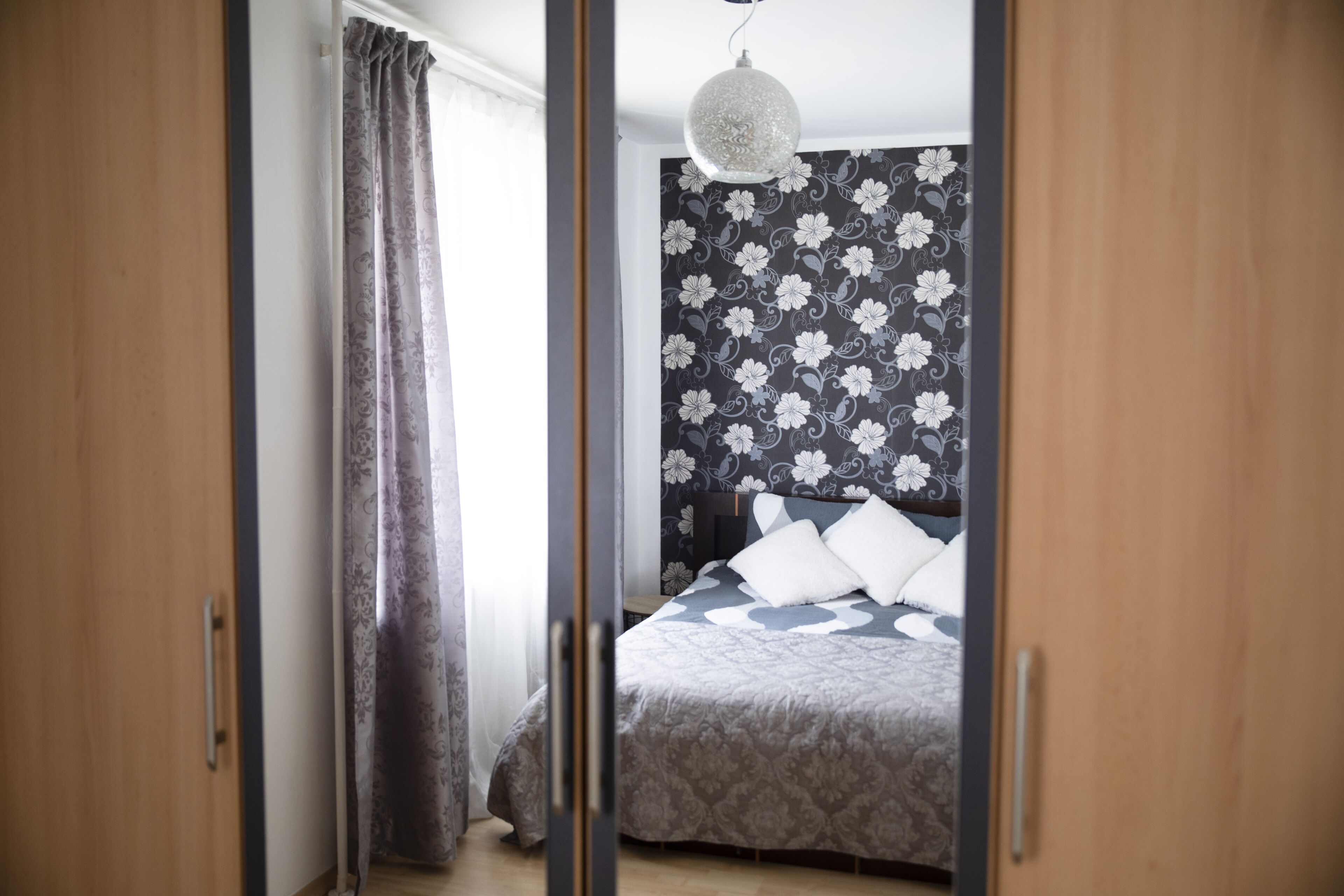 A bedroom with a bed, curtains, a hanging light fixture, and a floral-patterned wall.