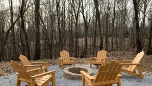 Een cirkel van houten Adirondack-stoelen rond een vuurkorf van steen, gelegen in een bosrijke omgeving.