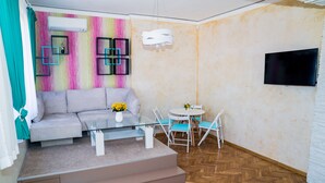 Ein modernes Wohnzimmer mit grauem Sofa, einem Glastisch und einer kleinen Essecke mit weißem Tisch und blauen Stühlen.