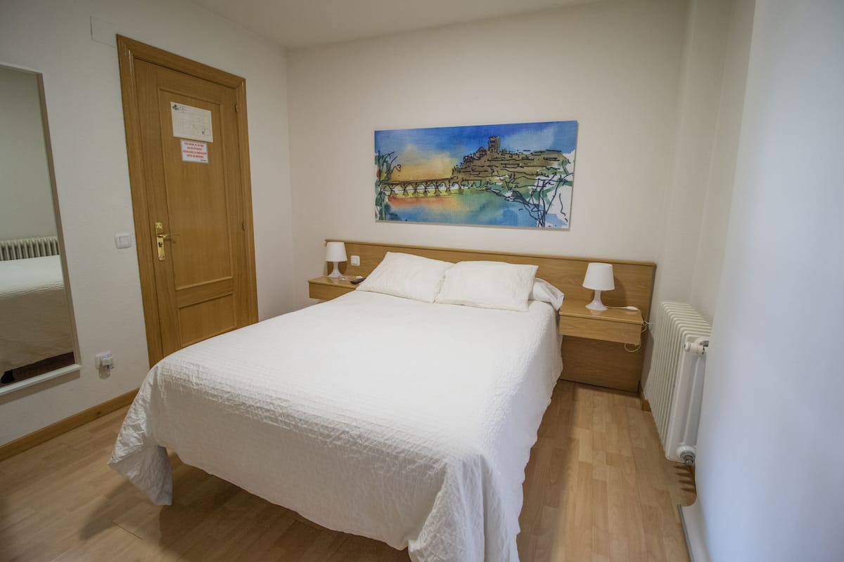 Un dormitorio con una cama, una mesita de noche, una lámpara, un cuadro en la pared y un radiador.