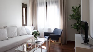 Una sala de estar moderna con un sofá, una mesa de centro, una silla, un televisor y un amplio ventanal con cortinas.