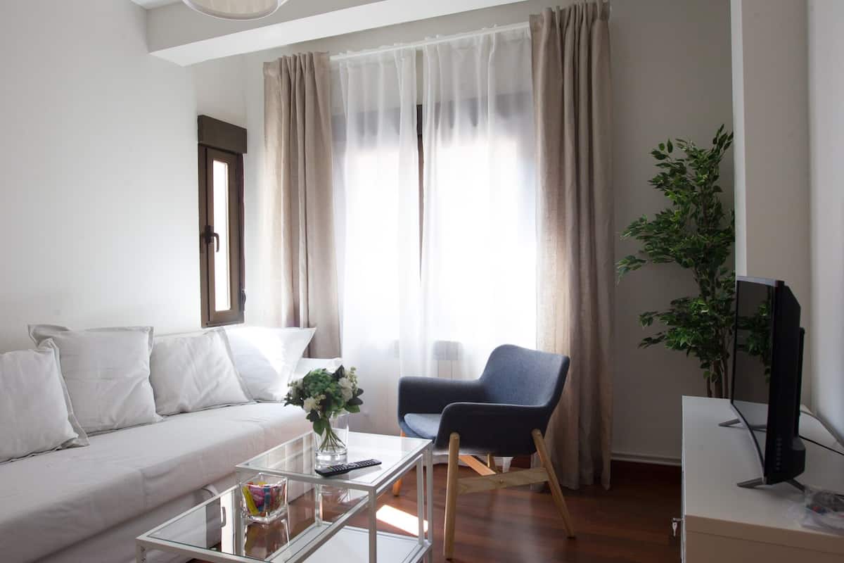 Una sala de estar moderna con un sofá, una mesa de centro, una silla, un televisor y un amplio ventanal con cortinas.