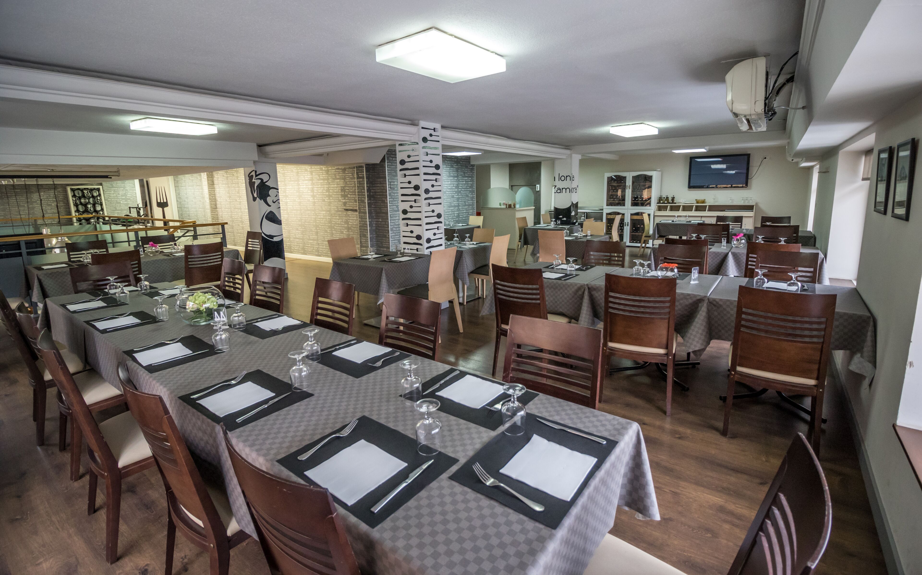 Un comedor con mesas dispuestas para el servicio, sillas de madera y una cocina al fondo.