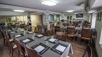 Un comedor con mesas dispuestas para el servicio, sillas de madera y una cocina al fondo.