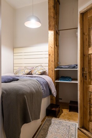 Un dormitorio con cama, una lámpara colgante, una viga de madera, un estante con toallas dobladas y una puerta.