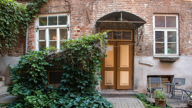 A brick building with a wooden door, a window, and a small outdoor seating area with metal chairs and a table.