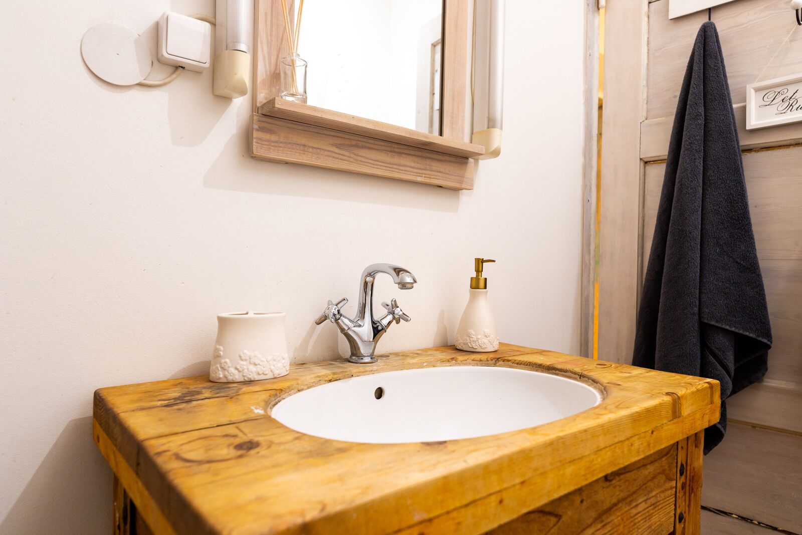 A bathroom with a wooden vanity, a white sink, a silver faucet, a small white vase, and a towel rack with a dark towel.