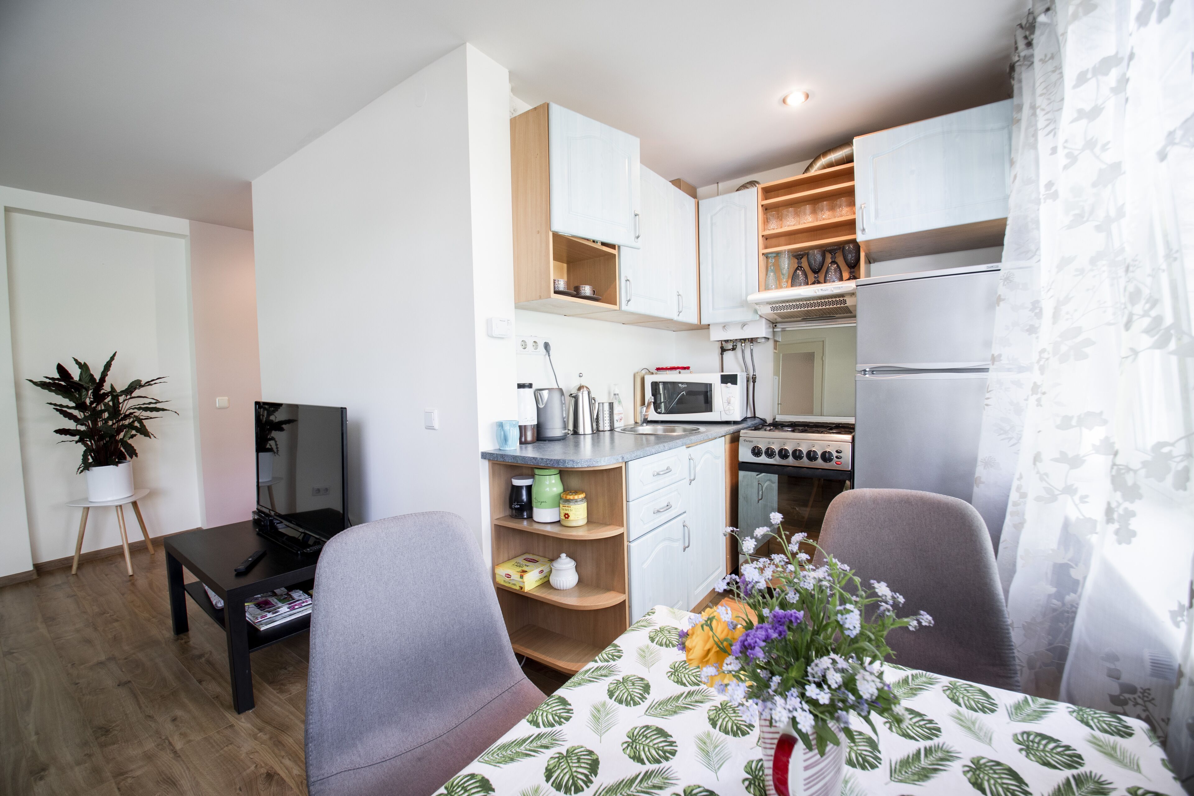 A compact kitchen with white cabinets, a microwave, and a stove. There is a dining table with a floral tablecloth and chairs. A potted plant is on a small table to the left.