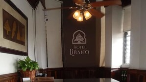 A reception desk with a logo for Hotel Libano, a ceiling fan, and a framed picture on the wall. - Hotel Libano (San Francisco de Macorís)