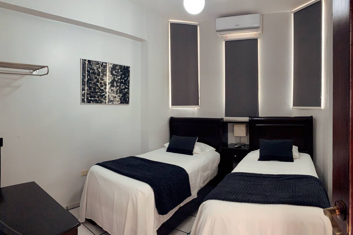 A hotel room with two beds, a desk, a wall-mounted air conditioner, and a modern abstract painting.