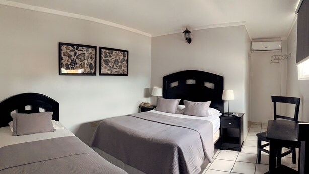 A hotel room with two beds, a nightstand with a lamp, a chair, and artwork on the wall. - Hotel Libano (San Francisco de Macorís)
