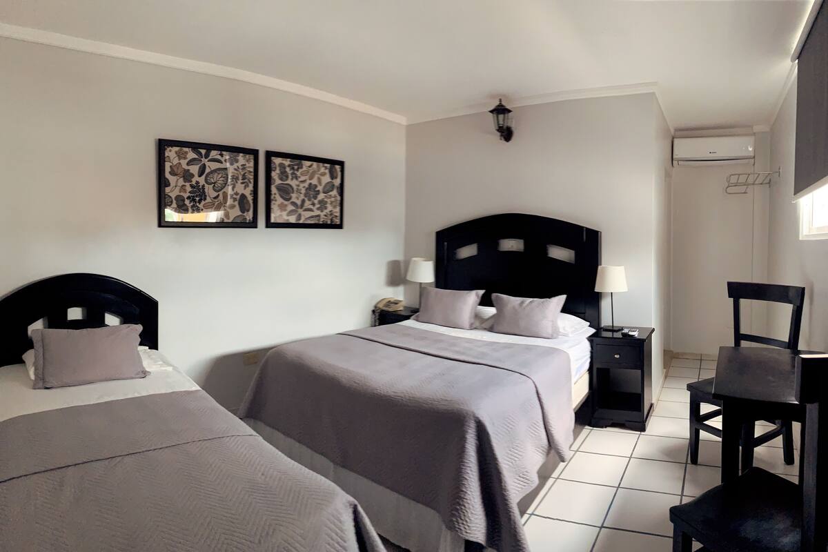 A hotel room with two beds, a nightstand with a lamp, a chair, and artwork on the wall.