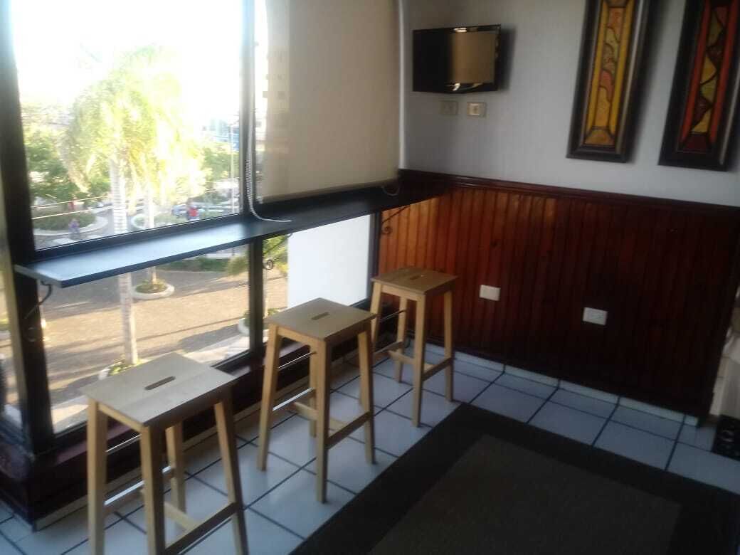 A room with a large window, wooden stools, a television mounted on the wall, and a framed picture.