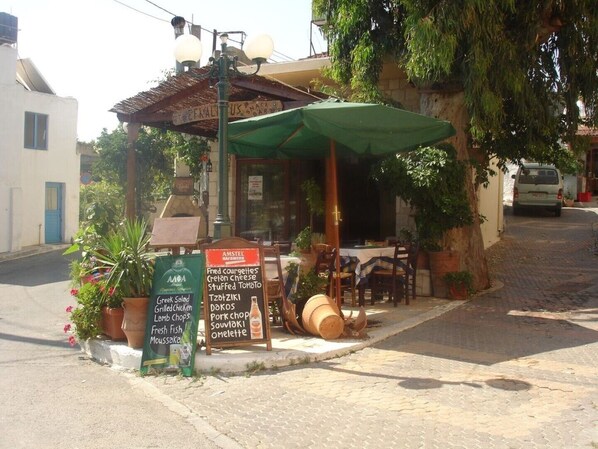A small outdoor café with a rustic wooden table and chairs, a green umbrella, and a menu board listing various dishes.
