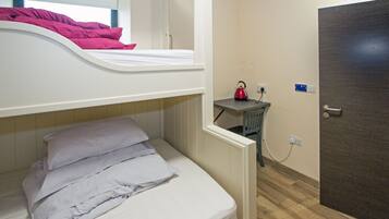 Una cama con literas, con una colcha rosa y una cama inferior con almohadas dobladas. Hay un hervidor rojo sobre una mesita de noche junto a un enchufe.