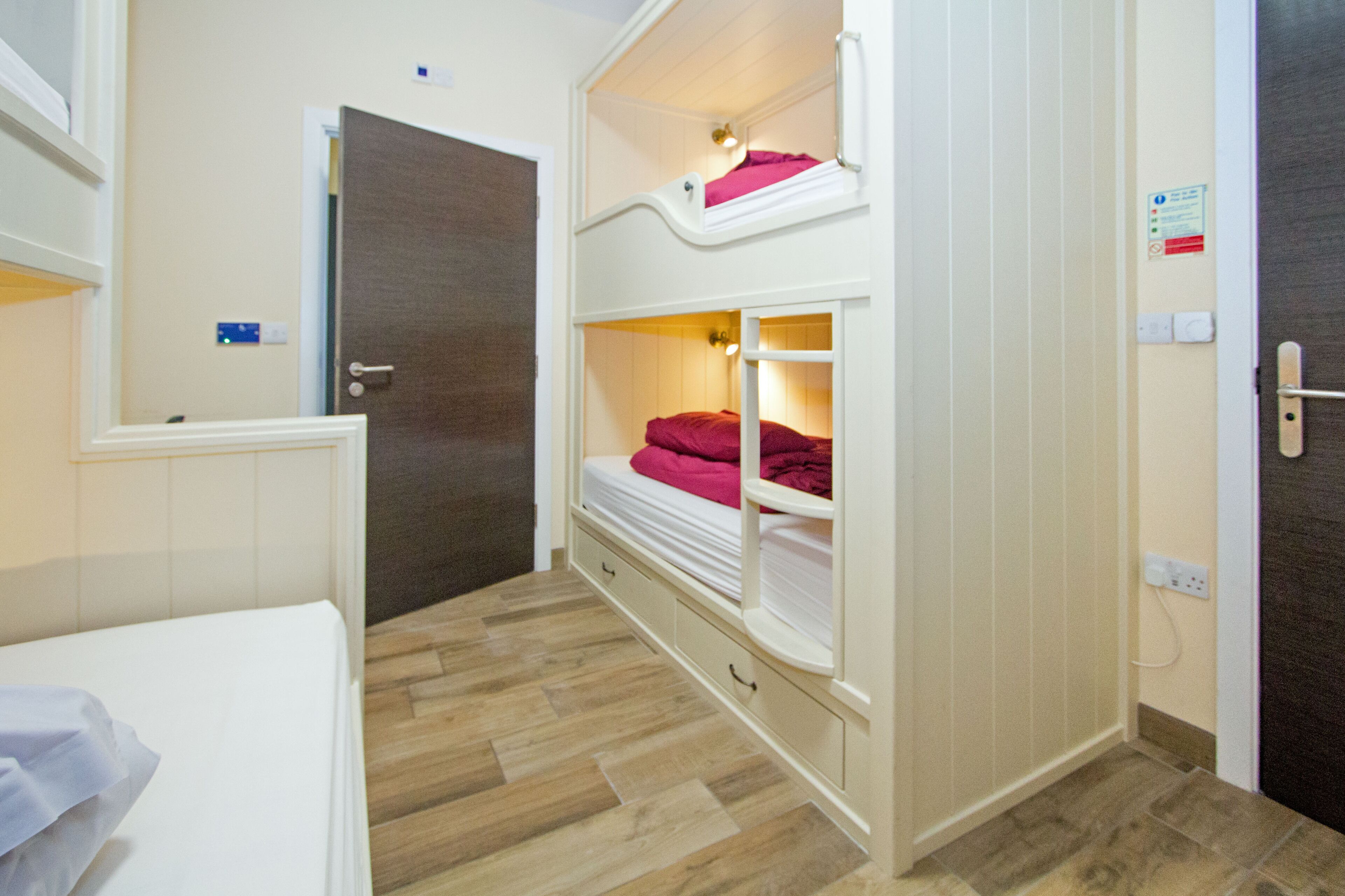 A bunk bed room with a wooden floor, a white door, and a small bedside table.