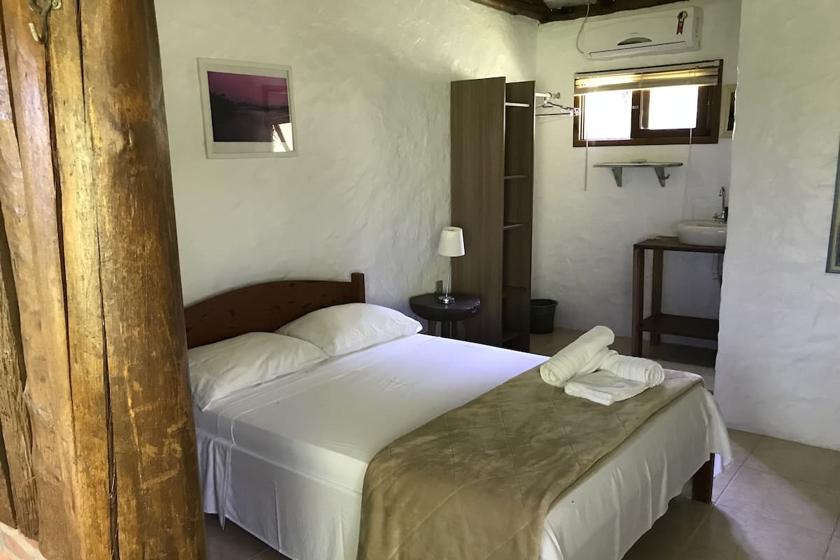 Ein Schlafzimmer mit Bett, Nachttisch mit Lampe, Holzschrank, kleines Badezimmer mit Waschbecken und Fenster mit Jalousien.