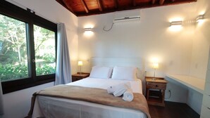 A neatly made bed with a beige blanket and white pillows, two bedside lamps, a small wooden nightstand, and a window with curtains. - Pousada Paraíso do Luz (Imbituba)