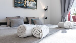 Cama bem arrumada com toalhas brancas dobradas, manta cinza e vários travesseiros.