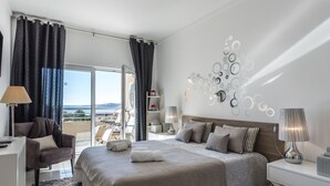 Quarto moderno com uma cama grande, duas mesas de cabeceira com luminárias, uma cadeira, vista para o exterior e um objeto decorativo na parede.