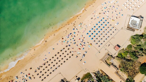 Vista aérea de uma praia com fileiras de guarda-sóis e espreguiçadeiras, um edifício com telhado vermelho e uma vista ampla do oceano.
