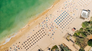 An aerial view of a beach with rows of umbrellas and sun loungers, a building with a red roof, and a clear view of the ocean.