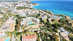 Vista aérea de um resort costeiro com vários edifícios, incluindo um hotel grande com piscina e um prédio menor com quadra de tênis.