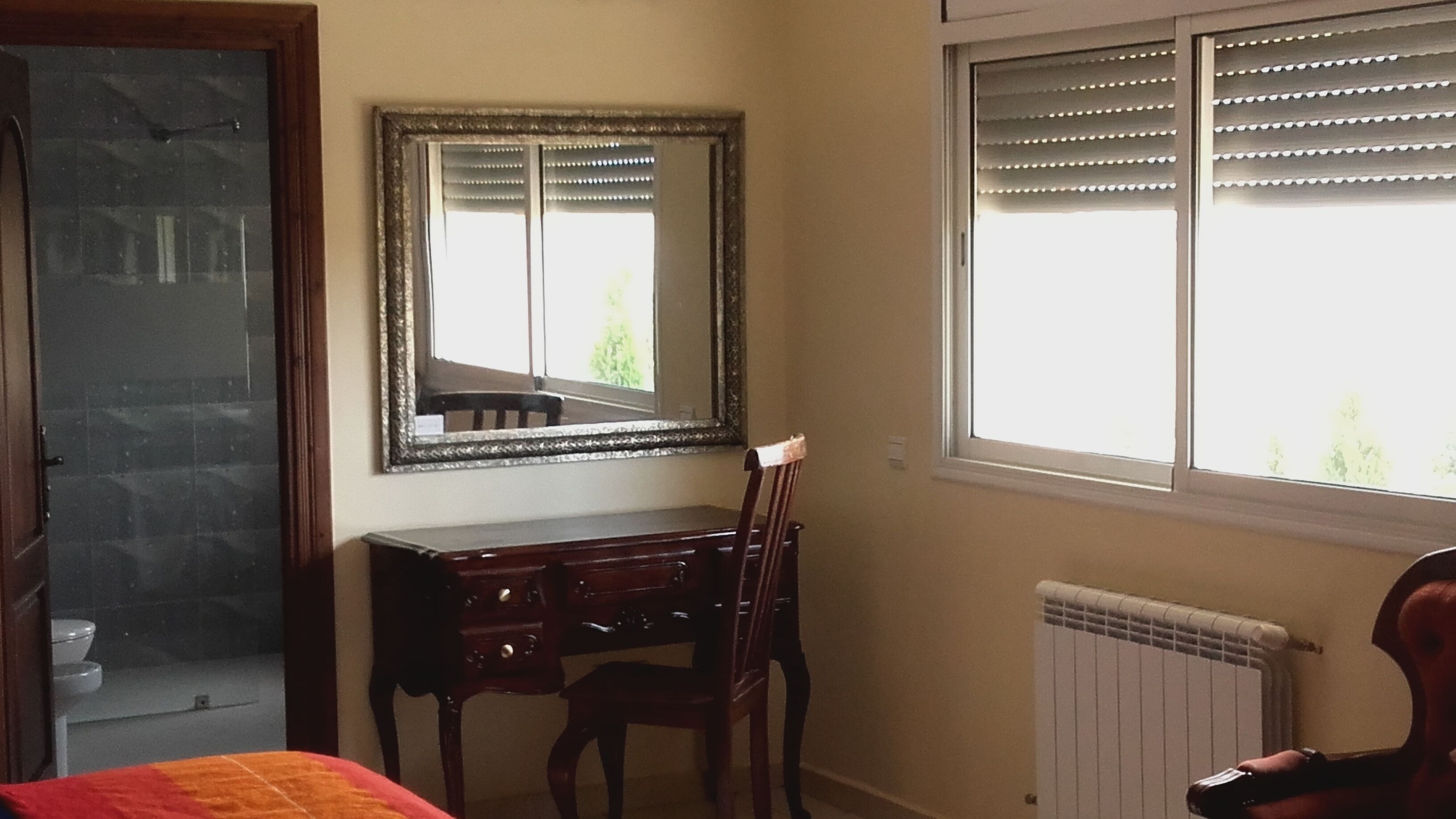 A room with a wooden desk, a chair, a large mirror, a radiator, and a window with blinds.