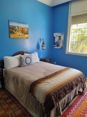 A bedroom with a bed, a nightstand, a wall-mounted lamp, and two paintings. - Riad Tanjil (Oulad Salah)