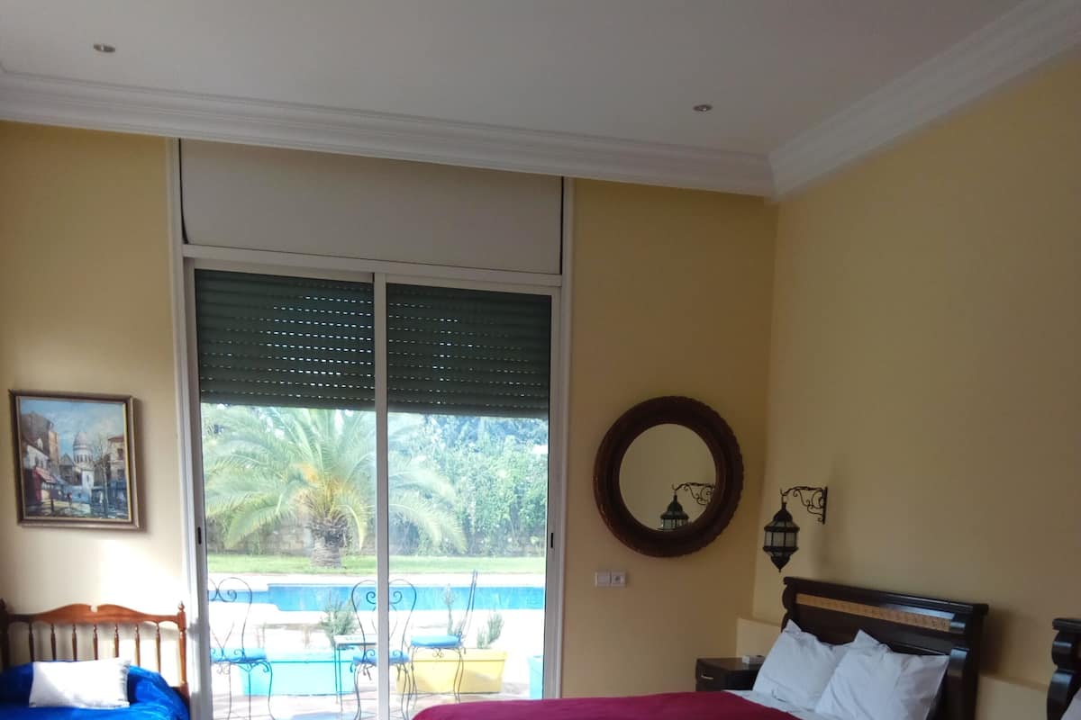Ett hotellrum med två sängar, en soffa, en mönstrad matta, en balkong med pool och utsikt över grönområden.
