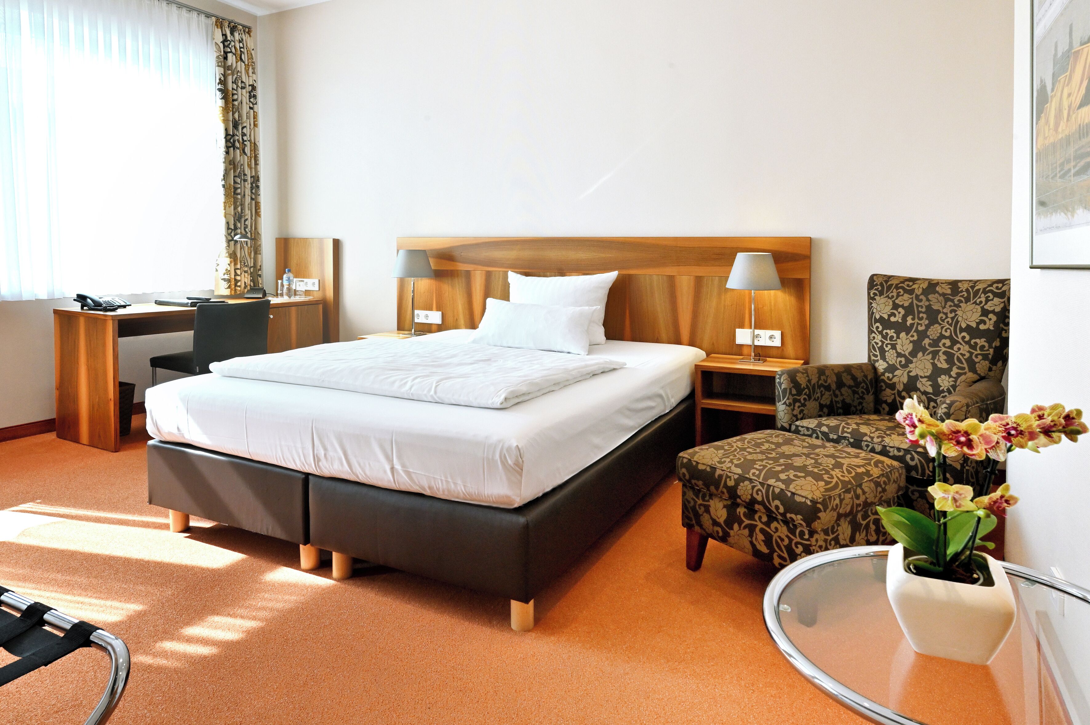 A hotel room with a bed, bedside tables, a desk, a chair, and a small table with a vase of flowers.