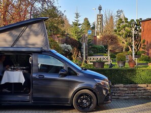A compact, modern vehicle with a covered cargo area, parked in a well-maintained garden with a 'HOTEL' sign in the background.