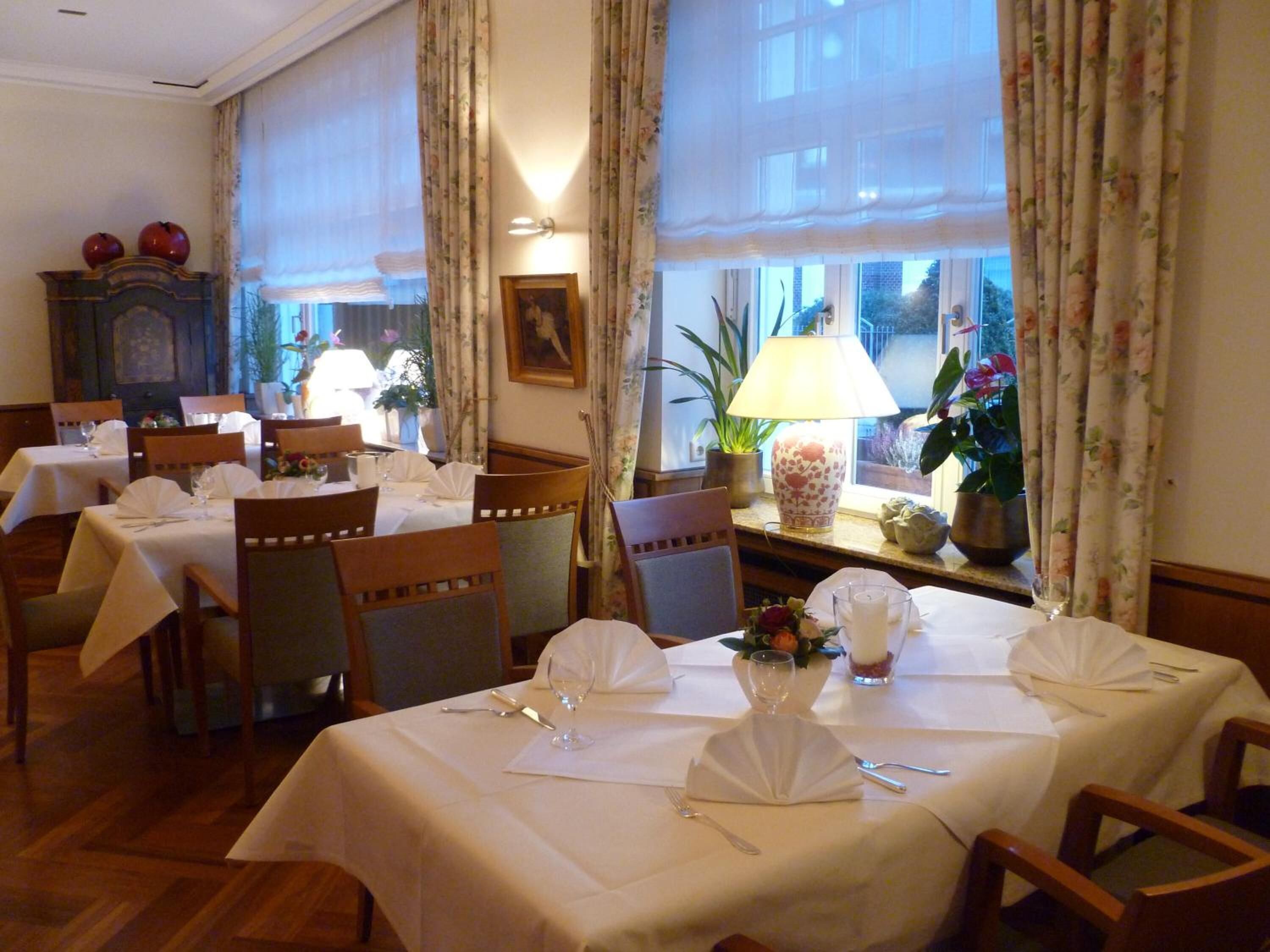 A dining area with tables set for two, wooden chairs, and a view of the outside through large windows.
