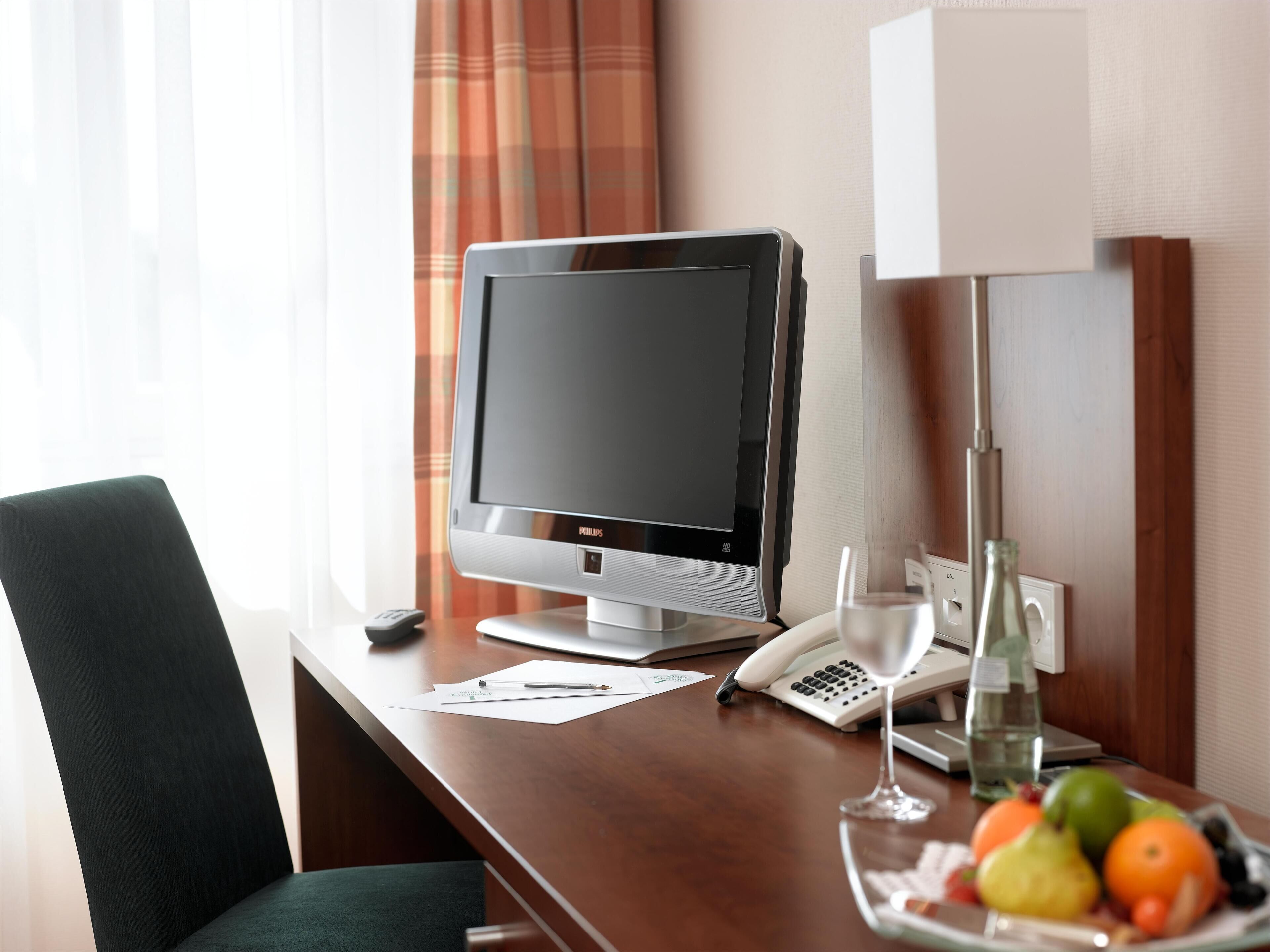 A hotel room with a desk, a flat-screen TV, a telephone, a glass of water, and a fruit bowl.