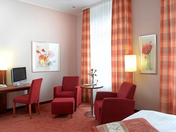 A hotel room with a bed, two red armchairs, a desk with a computer, a small table with a lamp, and two framed artworks on the wall.
