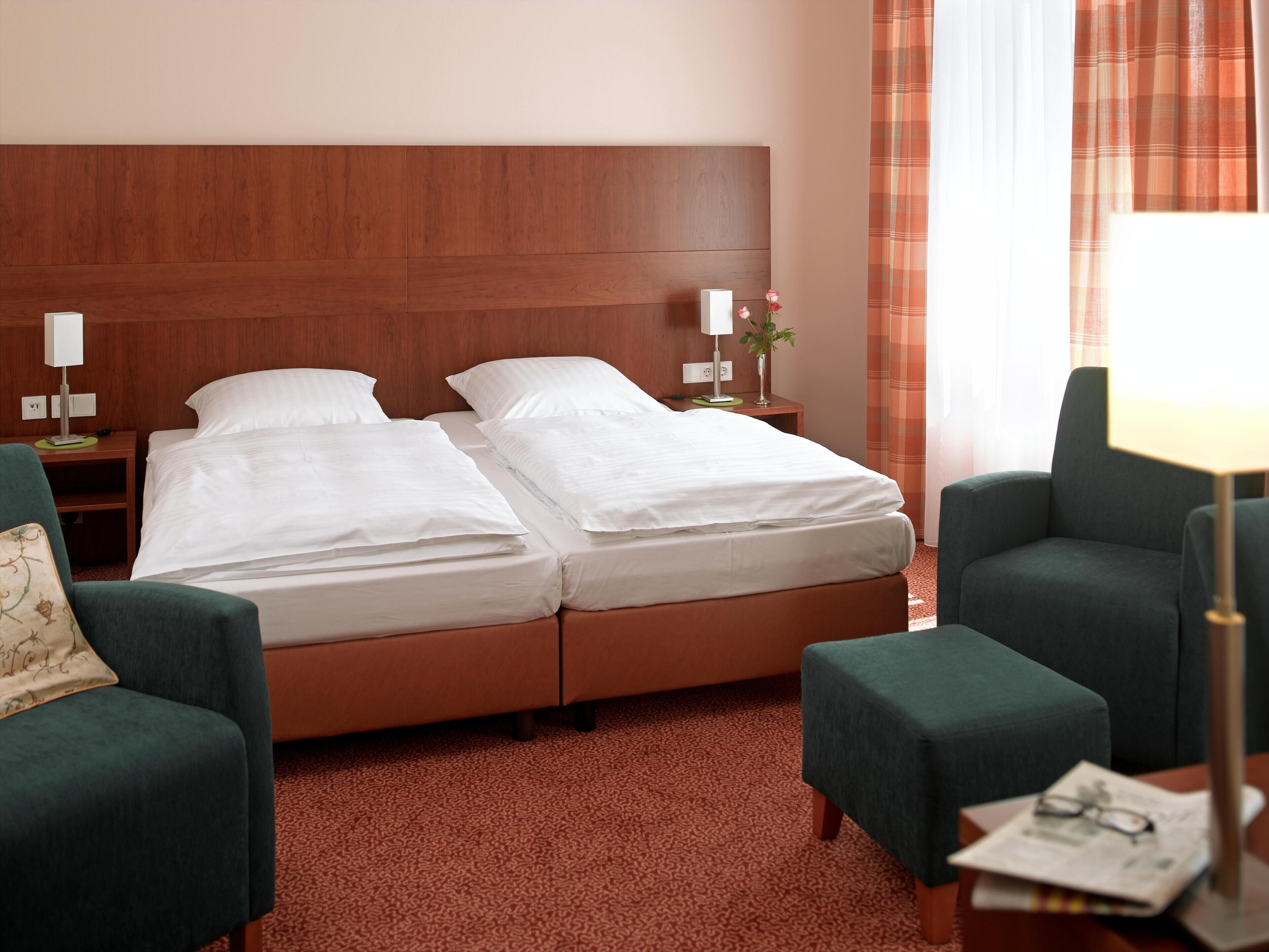 Ein Hotelzimmer mit zwei Betten, einem Holzkopfende, einem grünen Sessel, einem kleinen Tisch mit Lampe und einem Fenster mit Vorhängen.