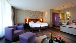 A hotel room with a bed, two chairs, a small table with a bottle and flowers, a bathroom with a sink and mirror, and a shower area. - Hotel Restaurant Lindenhof (Emsdetten)