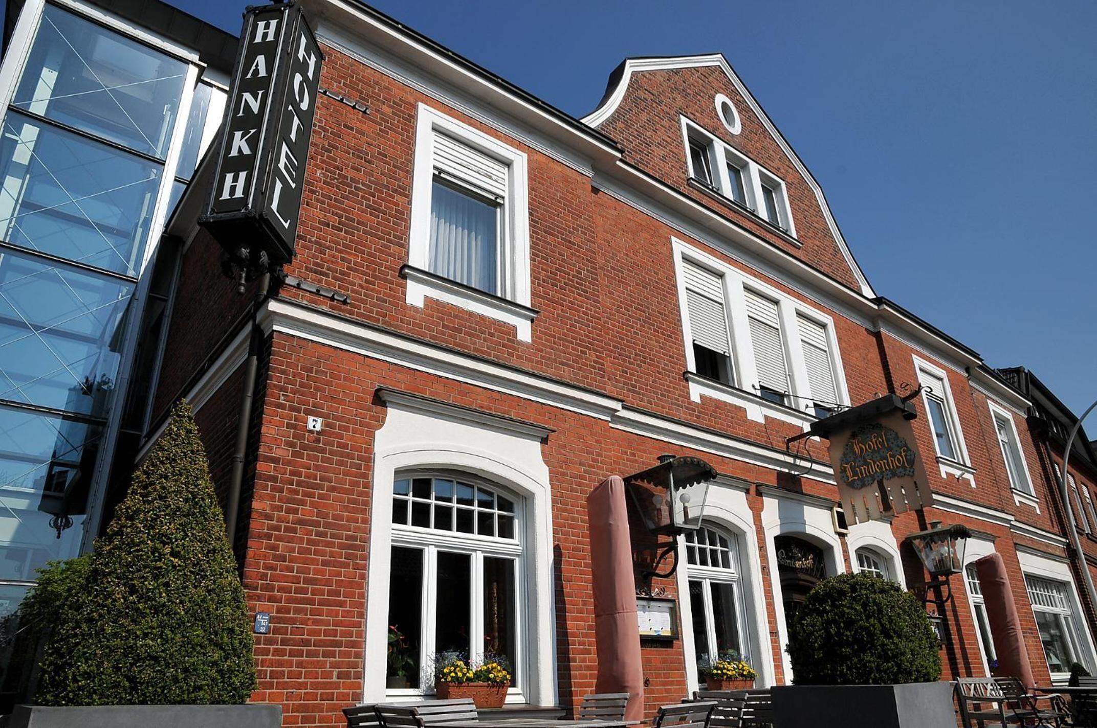 A red brick building with a sign reading "Hotel Lindenhof." It features large windows with white shutters, a prominent entrance with a canopy, and a small garden area with potted plants.